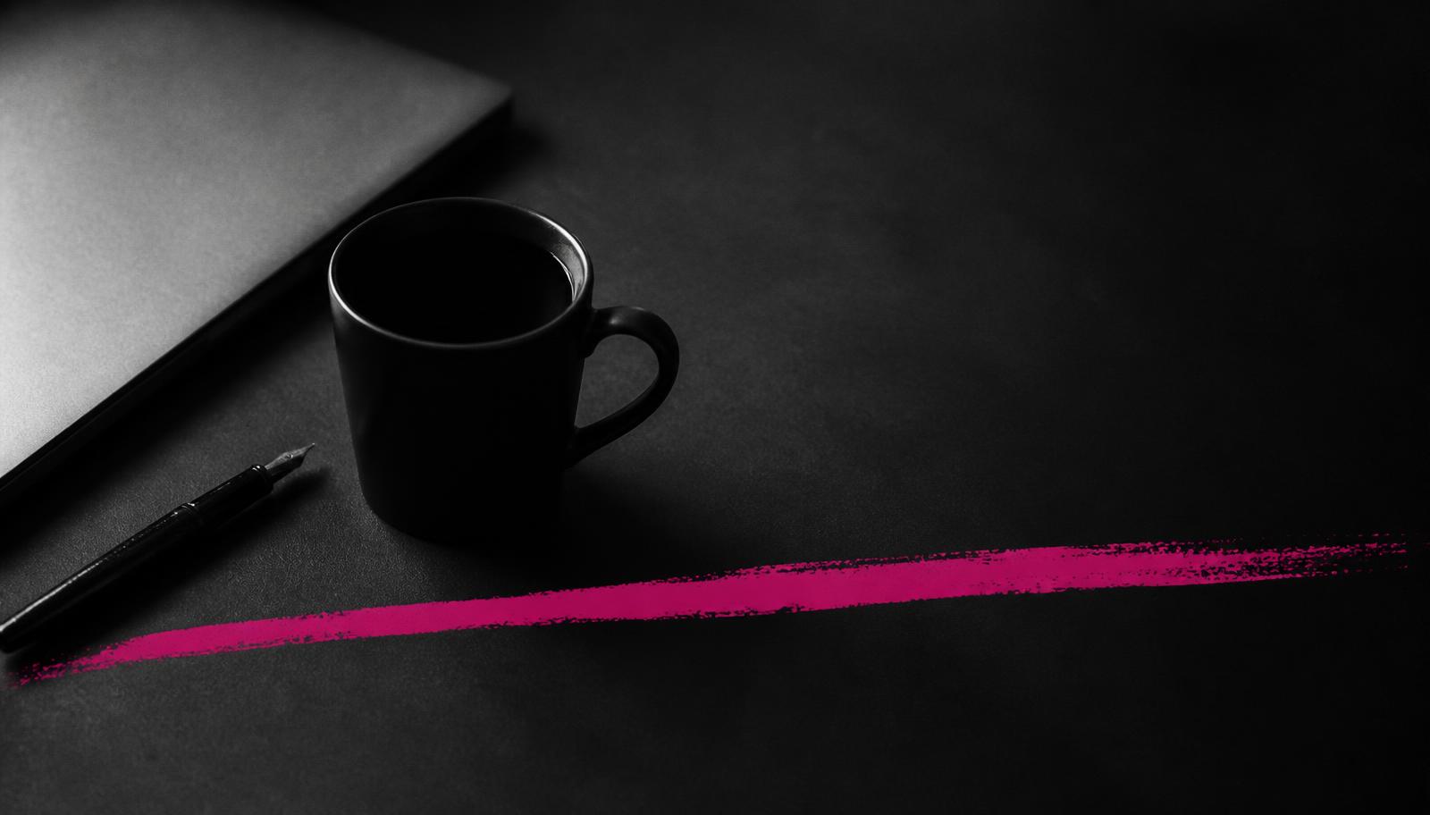 Black coffee mug, closed laptop and pen on a dark desk in black and white, cut by a single magenta brush stroke.
